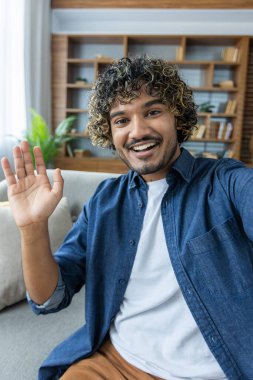 Dikey çekim, gülümseyen ve kameraya bakan adam, el sallayan merhaba, telefondaki uygulamayı video görüşmesi için kullanan adam. Evde, apartmanın oturma odasında oturmak..