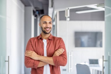 Genç işadamı, modern ofis ortamında kolları çapraz dururken kendinden emin bir şekilde gülümsüyor ve profesyonellik yayıyor.