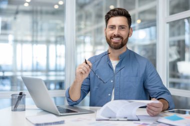 İş yerindeki başarılı finansör portresi, gülümseyen ve kameraya bakan adam. Grafikleri ve çizelgeleri olan, dizüstü bilgisayar kullanan bir iş adamı. Memnun muhasebeci yatırımcı.