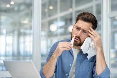 Elinde mendil ve burun spreyi olan hasta bir ofis çalışanı. Grip ve soğuk algınlığı olan bir adam. Ofisin içinde oturuyor..