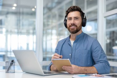 Bir adam, parlak ve modern bir ofis ortamında masanın üzerinde dizüstü bilgisayarla bir web sitesini izlerken ve notlar alırken görülüyor.
