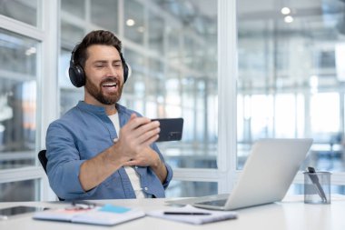 Kulaklıklı bir adam telefonuyla video izlerken gülüyor, masasında dinleniyor.