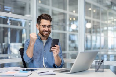 Ellerinde telefonla başarılı bir adam zaferi kutluyor ofis içinde iş yerinde. Bilgisayarla çalışan iş adamı, akıllı telefondan online uyarı aldı.
