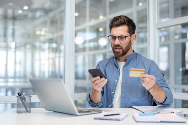 Masasındaki hayal kırıklığına uğramış bir adam, kredi kartı ve akıllı telefonuyla internet üzerinden ödeme yapmaya çalışırken bir hata fark ediyor.