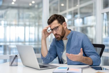 Kaygılı bir adam elinde dizüstü bilgisayarla masada oturuyor. Kötü haberlere ya da iyi bir ofis ortamında stresli bir duruma tepki veriyor.