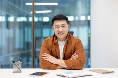 Asyalı işadamı modern bir ofiste masasında oturuyor. Kollarını kavuşturup kameraya gülümsüyor.
