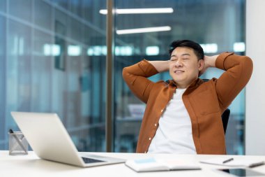 Ofisin içinde dinlenen ve dinlenen bir adam. Tatmin olmuş Asyalı adam ellerini kapalı gözlerle gülümseyerek başının arkasına koy. Başarılı sonuç, aferin..