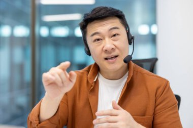 Webcam görüntüsü, kulaklıklı adam kameraya bakıyor, konuşuyor ve müşterilere uzaktan danışıyor. İş yerindeki destek servisi çalışanı ofis içinde, iş konferansı için görüntülü arama kullanıyor..