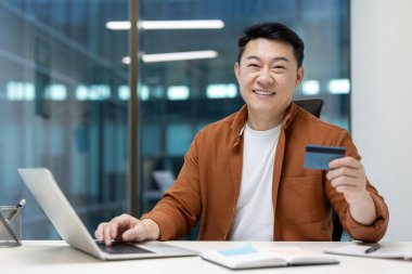 İş yerindeki mutlu ve başarılı Asyalı adamın portresi, banka kartı tutan adam, online alışveriş için dizüstü bilgisayar kullanan ve para transfer eden adam..