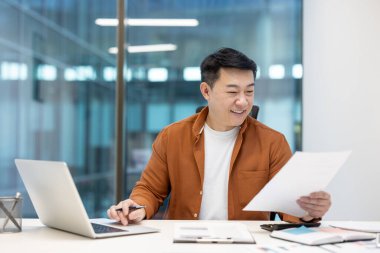 Başarılı bir iş adamı evrak işlerinde başarılı, Asyalı bir adam evrak raporları ve sözleşmelerle çalışıyor. Ofis içi bilgisayarı kullanan sıradan giysili bir adam..