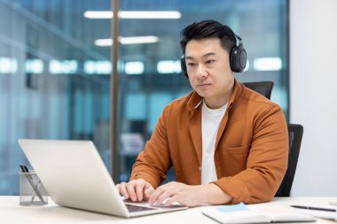 Ciddi özgüvenli ve odaklanmış Asyalı bir adam, ofiste dizüstü bilgisayarla çalışıyor, kulaklık takıyor, iş yerinde erkek programcı, yeni yazılım kodluyor..