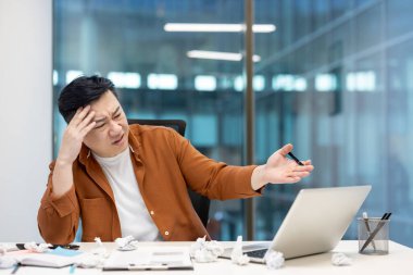 Hüsrana uğramış Asyalı işadamı, laptopunda çözüm arayan buruşuk kağıt toplarıyla çevrili bir şekilde ofiste tükenme yaşıyor.