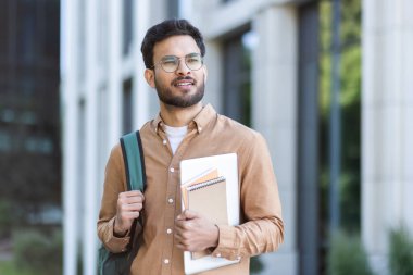 Üniversite kampüsünün dışında elinde kitaplarla dolaşan bir öğrenci. Sırt çantalı genç adam, mutlu ve gülümsüyor..