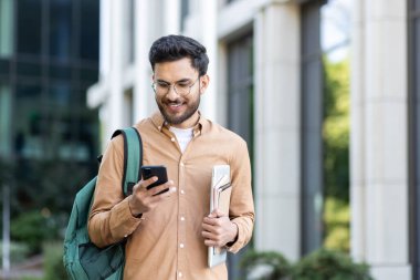 Sırt çantalı genç bir öğrenci akıllı telefonunda uygulama kullanıyor. Üniversite kampüsünün dışında elinde telefonla gülümseyen bir adam..
