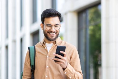 Sırt çantalı genç bir öğrenci akıllı telefonunda uygulama kullanıyor. Üniversite kampüsünün dışında elinde telefonla gülümseyen bir adam..