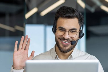 Ofis içinde kulaklığı ve dizüstü bilgisayarı olan destek servisi çalışanı. Selamlaşmak için el sallayan adam. Danışmanlık ve müşterilere yardım için video çağrısı kullanılıyor.