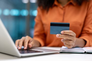 Bir kadın dizüstü bilgisayarda yazarken elinde kredi kartı tutuyor, görünüşe göre online alışveriş yapıyor ya da mali durumunu yönetiyor.