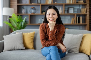 Asyalı bir kadın rahat bir kanepeye oturmuş, günlük kahverengi bir polka bluz ve kot pantolon giyiyor. Ev dekoru ve kitaplıklarla çevrili rahat ve neşeli bir ifade..