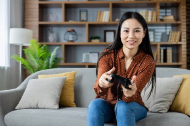 Kanepede oturan Asyalı kadın kontrolörle video oyunu oynuyor. Rahatlamış ve gülümsüyor, evde eğleniyor. Rahat, modern oturma odası düzenleme eğlenceli oyun deneyimi taşır.