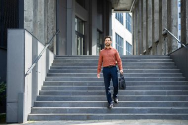 Kendinden emin bir işadamı, güneşli bir günde çantasıyla modern bir ofis binasının merdivenlerinden iniyor. Yavaşça gülümsüyor.