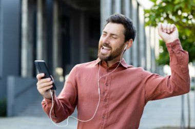 Neşeli bir adam akıllı telefonuyla kulaklıkla müzik dinliyor, ritim ve açık hava müziğinin tadını çıkarıyor.