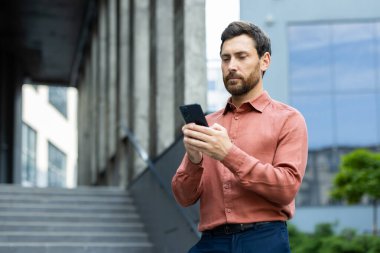 Gömlekli yakışıklı bir adam modern bir binanın dışında akıllı telefonunu kontrol ediyor, muhtemelen iş arkadaşları ya da müşterileriyle iletişim kuruyor.