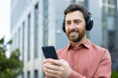 Gülümseyen mutlu adam kulaklık takıyor, akıllı telefondaki uygulamayı kullanarak müzik dinliyor. İş adamı elinde telefonla ofis binasının önünde yürüyor, ses kitapları ve yayınları dinliyor..