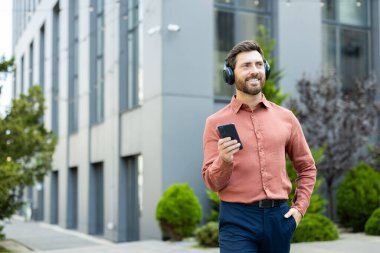Yakışıklı bir adam ofis binasının yanında yürüyor, akıllı telefonunu kullanarak kulaklıkla müzik dinliyor.