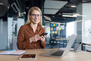 Ofiste başarılı bir kadının portresi, elinde telefonla kameraya bakıp gülümseyen bir iş kadını. Günlük kıyafetlerle çalışan şirket çalışanı masasında mutlu..