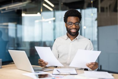 Gözlüklü, gülümseyen Afrikalı Amerikalı işadamı modern bir ofiste masasında dokümanları inceleyerek, olumlu bir çalışma ortamı sergiliyor.