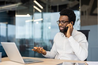 İşyerinde dizüstü bilgisayarla masada oturan başarılı bir adam telefonda konuşuyor. İş adamı, meslektaşları ve ortaklarıyla görüşmelere odaklanmış durumda..