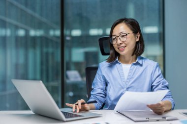 Ofiste çalışan bir iş kadını, gece geç saatlerde yüzünde bir gülümsemeyle dizüstü bilgisayarını kullanıyor ve belgeleri inceliyor..