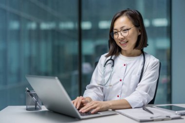 Ofisinde dizüstü bilgisayarla çalışan Asyalı bir doktor. O, gülümserken ve bilgisayarında yazarken beyaz bir önlük ve steteskop giyiyor.