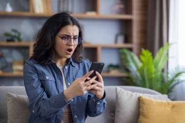 Genç bir kadın koltuğunda oturur ve elinde akıllı telefonuyla şoke olmuş bir şekilde bakar. İç mekan ayarları, sıradan bir kot gömlek giyer..