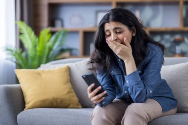 Bir kadın evdeki bir kanepede otururken telefonuna bakıp üzüntü ya da şok belirtileri gösteriyor..