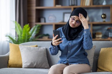 Başörtüsü içinde genç bir Müslüman kadın telefonunu evde kullanırken stresli görünüyor, muhtemelen kötü haber alıyor ya da bir sorunla uğraşıyor..