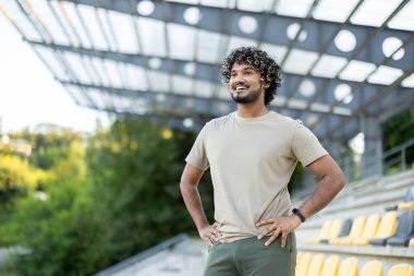 Genç erkek sporcu dinleniyor, elleri kalçasında, ayakta gülümsüyor ve nefes alıyor, aktif fiziksel egzersiz ve spor yaptıktan sonra dinleniyor..