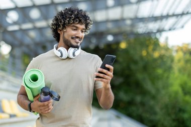 Genç bir adam telefonunda uygulama kullanıyor, bir atlet yoga minderiyle antrenmanlara gidiyor, mutlu bir şekilde gülümsüyor.