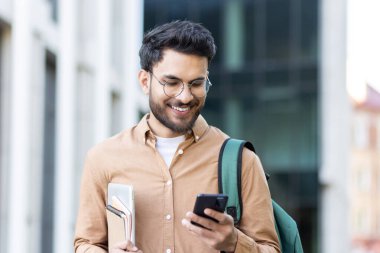 Sırt çantalı ve kitaplı gülümseyen öğrenci kampüsteyken cep telefonunu kontrol ediyor.