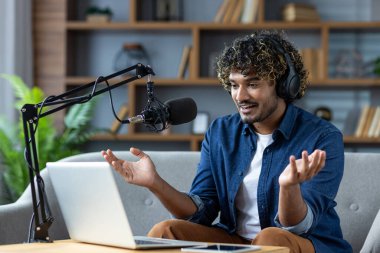 Genç adam heyecanla bir podcast kaydediyor ya da oturma odasından canlı bir online toplantıya katılıyor, mikrofon ve dizüstü bilgisayar kullanarak..
