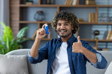 Kıvırcık saçlı, gülümseyen ve astım spreyini evde tutarken baş parmağını kaldıran genç etnik adam, solunum sağlığını destekliyor..