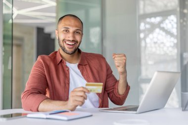 Banka kartı ve laptopuyla kameraya bakan neşeli ve başarılı bir adam. Şirket çalışanı başarılı online para transferini kutluyor.