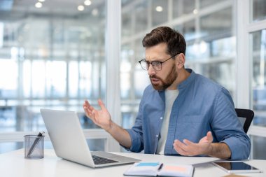Modern bir ofisteki bir işadamı dizüstü bilgisayarına bakarken hayal kırıklığı yaşar, muhtemelen bir sorun veya hatayla karşılaşır..