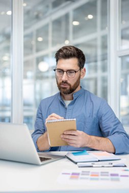 Çevrimiçi iş toplantısı, konferans. İş yerindeki adam video görüşmesi için dizüstü bilgisayar kullanıyor, not defterine veri yazıyor, ciddi ve konsantre bir şekilde iş arkadaşlarını dinliyor. Uzaktan çalışmak, webinar izlemek..