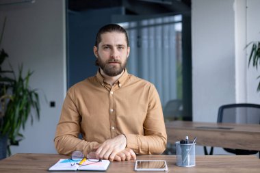 Sakallı bir adam masasında tablet ve not defteriyle modern ofis ortamında oturuyor. Ciddi bir bakışı var ve önüne bakıyor..