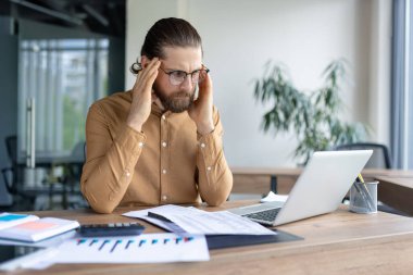 Ofisindeki dizüstü bilgisayara bakarken şakaklarına masaj yapan stresli bir işadamı, haritalar ve mali belgelerle çevrili..