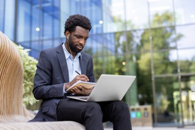 Takım elbiseli odaklanmış işadamı notlar yazarken, kulaklık takarken ve modern bir binanın arka planında çalışırken dışarıda dizüstü bilgisayarda çalışıyor..