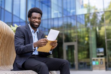 Gülümseyen bir iş adamı, içinde muhtemelen iyi haberler olan bir zarfı açarken, modern bir bankta ofis binasının önünde oturuyor..