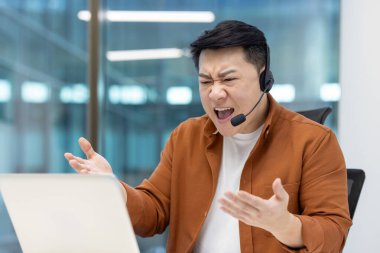 Kızgın bir Asyalı müşteri hizmetleri temsilcisi, modern bir ofis ortamında kulaklık takarken dizüstü bilgisayarına bağırıyor.