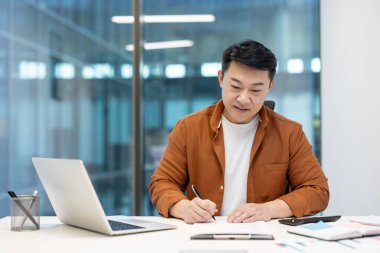 Başarılı muhasebeci evrak işlerinde başarılı, Asyalı adam gülerek vergi formlarını dolduruyor, rapor hazırlıyor. Belgeler ve faturalar arasında bir işadamı.